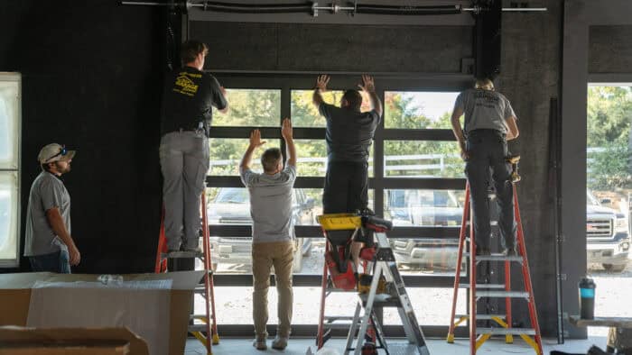 Five workers skillfully install a large commercial overhead door using ladders and tools inside a building.