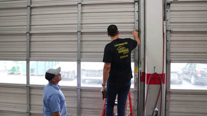 Two men are inspecting a garage door. One, standing on a ladder, holds a tool while the other, wearing a cap, observes from below. They discuss garage door repair options to ensure smooth operation and consider whether commercial overhead doors might be appropriate for their needs.
