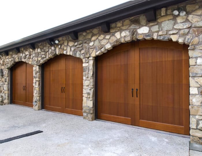 Stone facade with three large wooden garage doors, each featuring black handles, set on a textured concrete driveway.