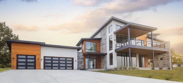Modern two-story house with a mix of wood, stone, and glass elements, featuring a large driveway and a double garage under a partly cloudy sky.