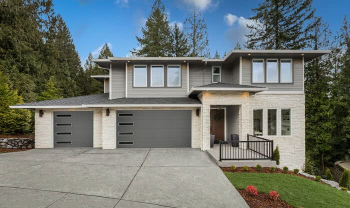 Modern two-story house with gray siding, large windows, and a three-car garage. Surrounded by tall trees, with a wide driveway and landscaped front yard.