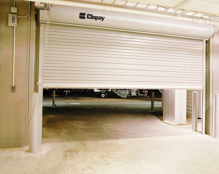 A partially opened commercial overhead door with Clopay branding offers a glimpse of the outside area.